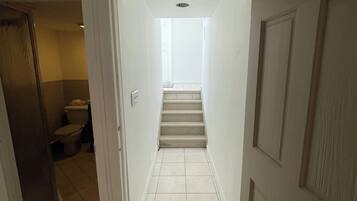 A narrow hallway with a staircase leading to an upper floor. The walls are white, and the floor is tiled.
