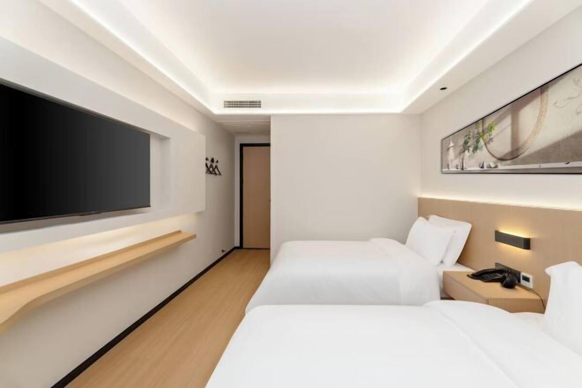 A modern hotel room with two beds, a flat-screen TV mounted on the wall, a bedside table with a lamp, and a painting above the bed.
