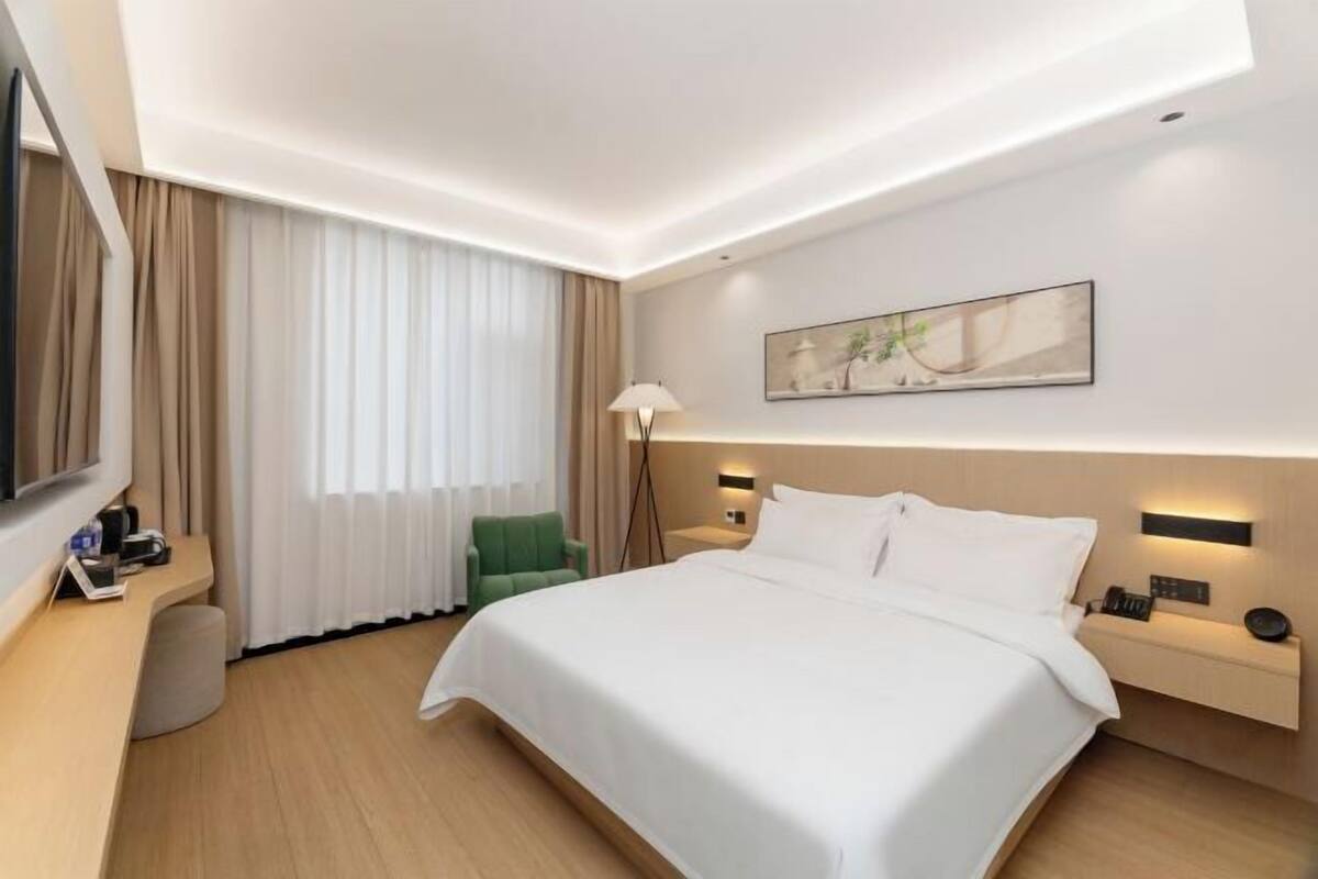 A modern hotel room with a large bed, a flat-screen TV, a desk with a chair, and a bedside table with a lamp.