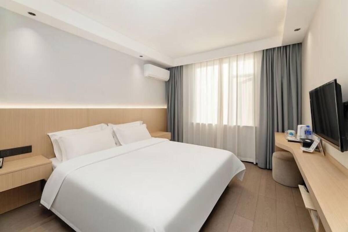 A modern hotel room with a large bed, a desk with a flat-screen TV, and a window with curtains.