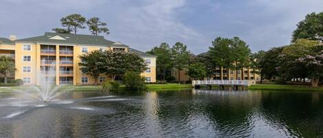 A multi-story building with balconies, a pond with a fountain, and a bridge.