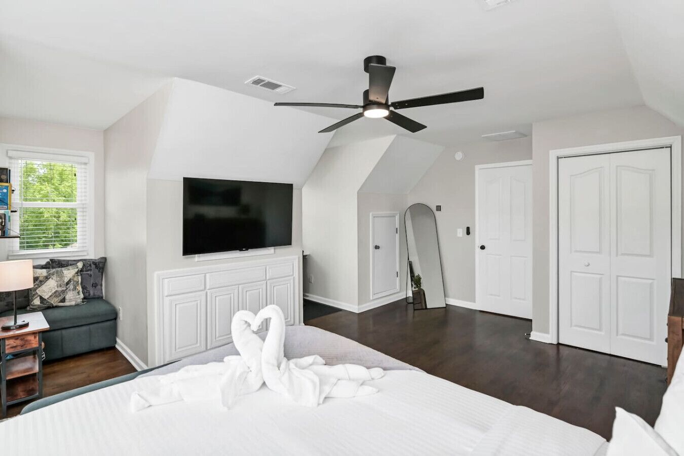 Un dormitorio con cama, televisor, ventilador de techo, espejo grande y puertas blancas.