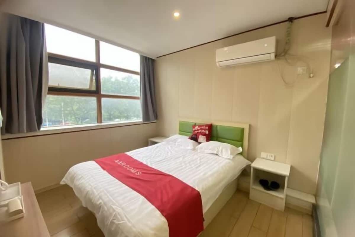Una habitación de hotel con una cama, una mesita de noche, una ventana con cortinas, un aire acondicionado y un estante con zapatos.