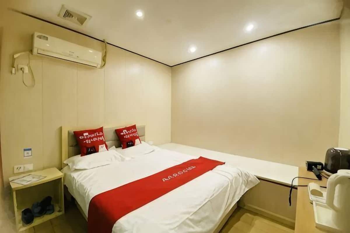 Habitación de hotel con cama, mesita de noche, una manta roja y una cafetera blanca.
