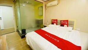 Une chambre d’hôtel avec un lit, une cabine de douche en verre, un mur blanc avec un appareil de climatisation et une couverture de lit rouge.