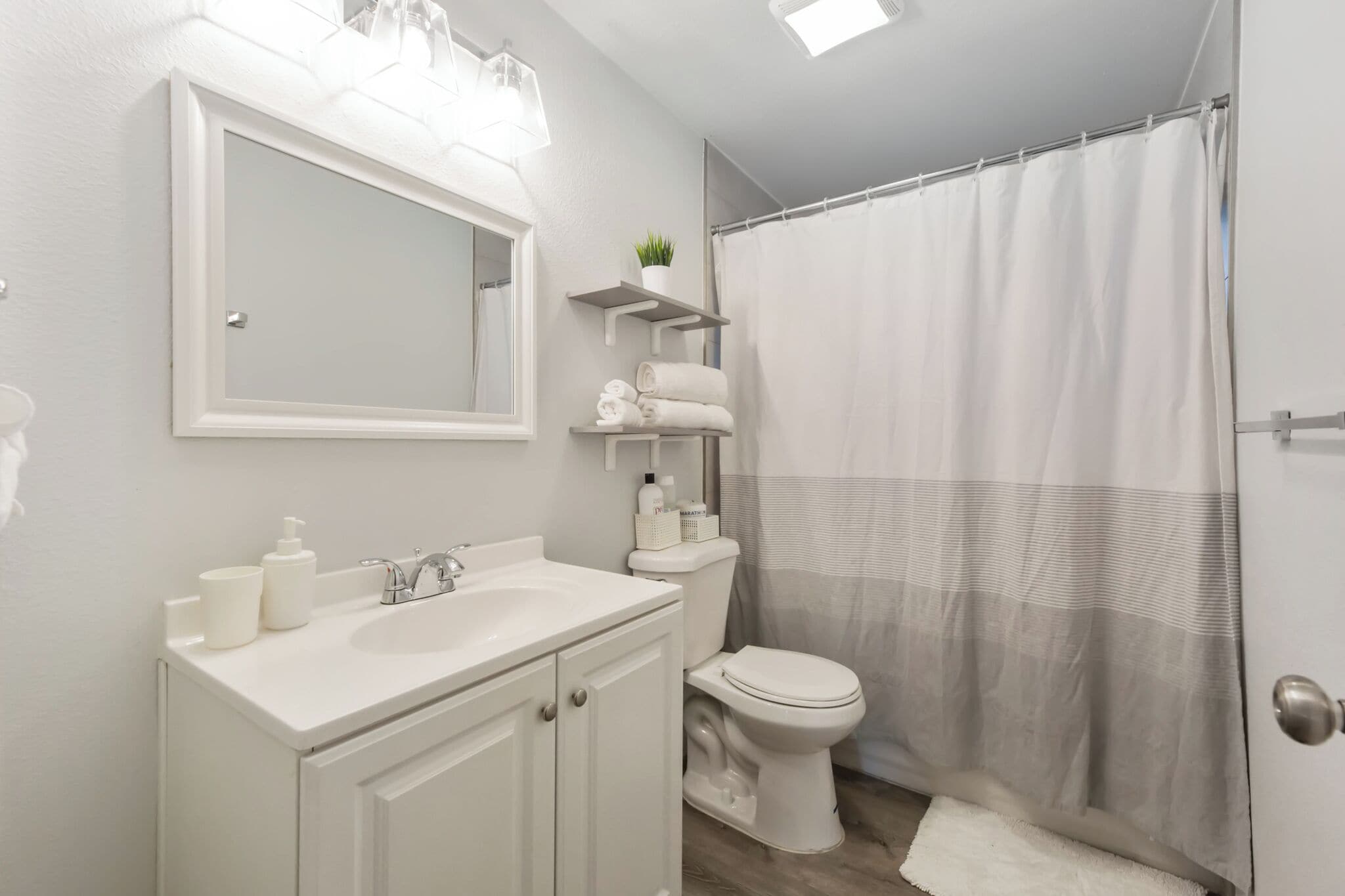 Une salle de bain avec un lavabo blanc, un miroir et des toilettes. On y trouve des étagères avec des serviettes et des articles de toilette.