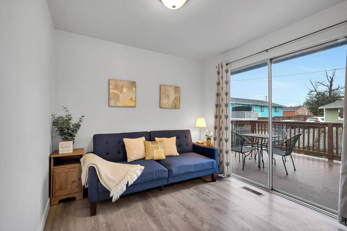 A living room with a blue sofa, two cushions, a small wooden side table with a plant, and a glass door leading to an outdoor patio with a table and chairs.