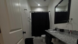 A narrow bathroom with a white door, a black trash bin, a shower curtain, a toilet, and a granite countertop with a mirror. - Gates at Sour Lake #502 (Sour Lake)
