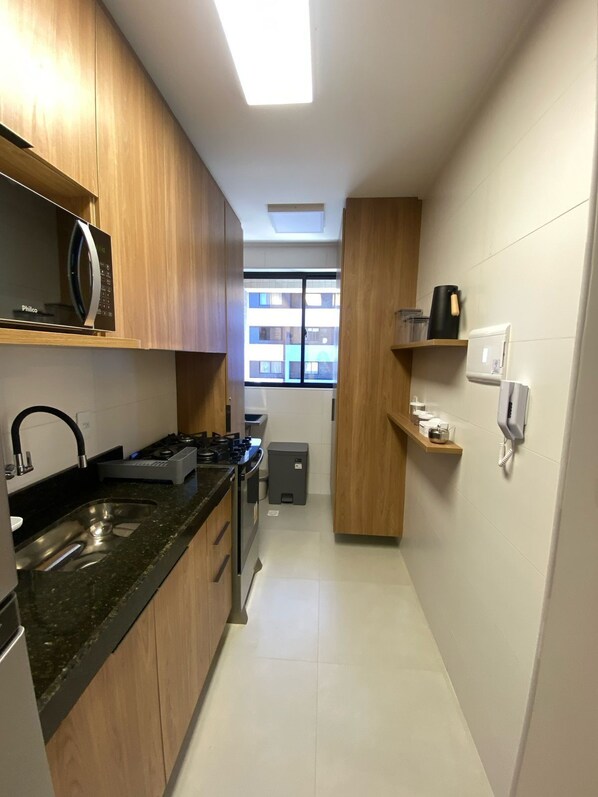 A narrow kitchen with wooden cabinets, a gas stove, a sink, and a window with a view of buildings. - Studio Design 3 Apto 810-Mandi  (Maceio)