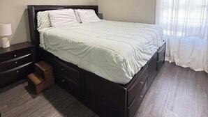 Een slaapkamer met een bed, nachtkastje, ladekast, plafondventilator en een raam met gordijnen.