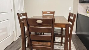 Een eetgedeelte met een houten tafel en stoelen, een vloerkleed met dessin, en een keuken met een donker aanrecht en kastjes.