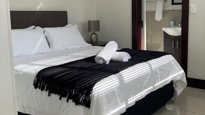 Ein ordentlich bezogenes Bett mit weißen Leinentüchern, einem dunklen Kopfteil und einer schwarz-weißen Streifendecke. Ein kleiner Nachttisch steht daneben, mit einer Lampe und einem gerahmten Bild an der Wand über dem Bett.