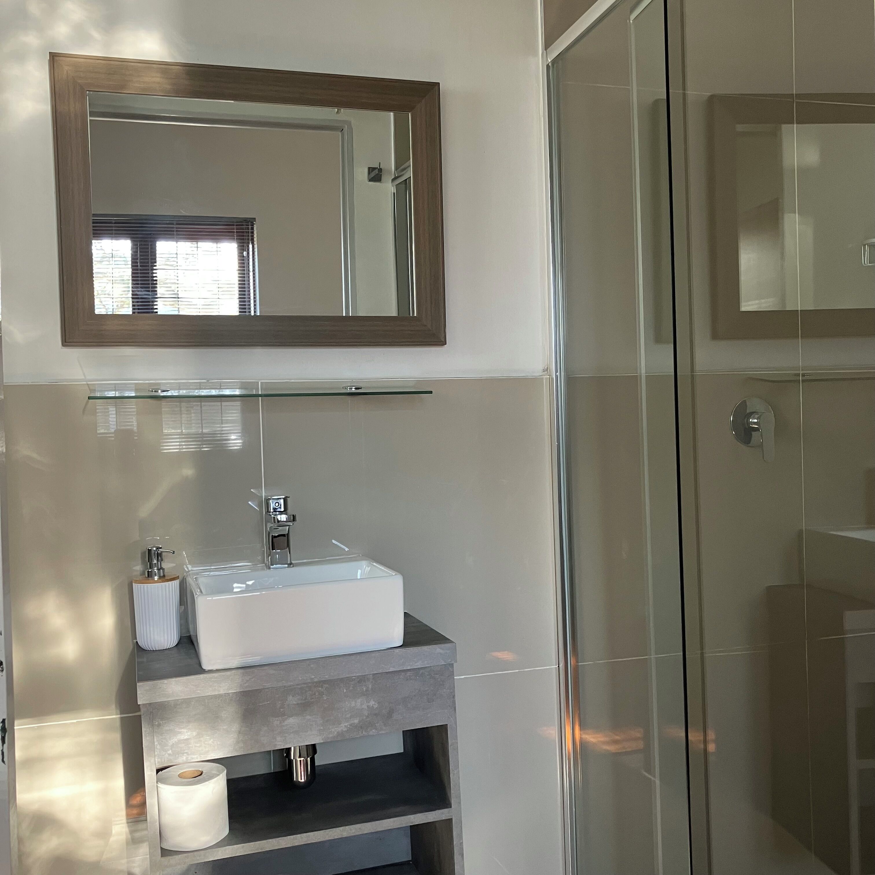 Un baño con cabina de ducha de vidrio, un lavabo blanco con estante de madera y un espejo sobre el lavabo.