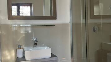 Un baño con cabina de ducha de vidrio, un lavabo blanco con estante de madera y un espejo sobre el lavabo.