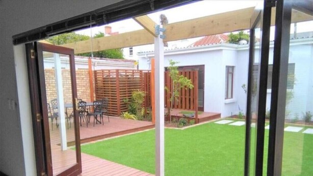 Blick auf eine Terrasse mit Holzdeck, einer kleinen Rasenfläche und einem weißen Gebäude im Hintergrund.