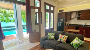 A modern living room with a grey sofa, patterned cushions, and a round wooden stool. A kitchen area with dark cabinets and a dining table is visible in the background. - Boutique Hotel Casa Grande, Parrot Paradise Suite, 1 BR (Santo Domingo)
