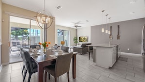A modern kitchen with a central island, a dining area with a chandelier, and a living space with a sofa and TV. - Relax Near Disney 4BR Home With Pool (Kissimmee)