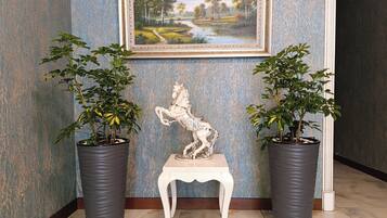 A hallway with a framed landscape painting, a white pedestal with a horse statue, and two potted plants on either side.
