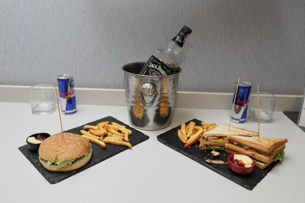 Una mesa con dos disposiciones de comida, incluyendo una hamburguesa, papas fritas y un sándwich, junto con bebidas y una botella de whisky Jack Daniel's.