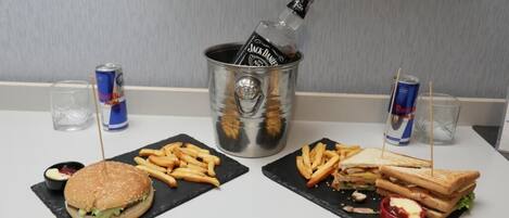 Una mesa con dos disposiciones de comida, incluyendo una hamburguesa, papas fritas y un sándwich, junto con bebidas y una botella de whisky Jack Daniel's.