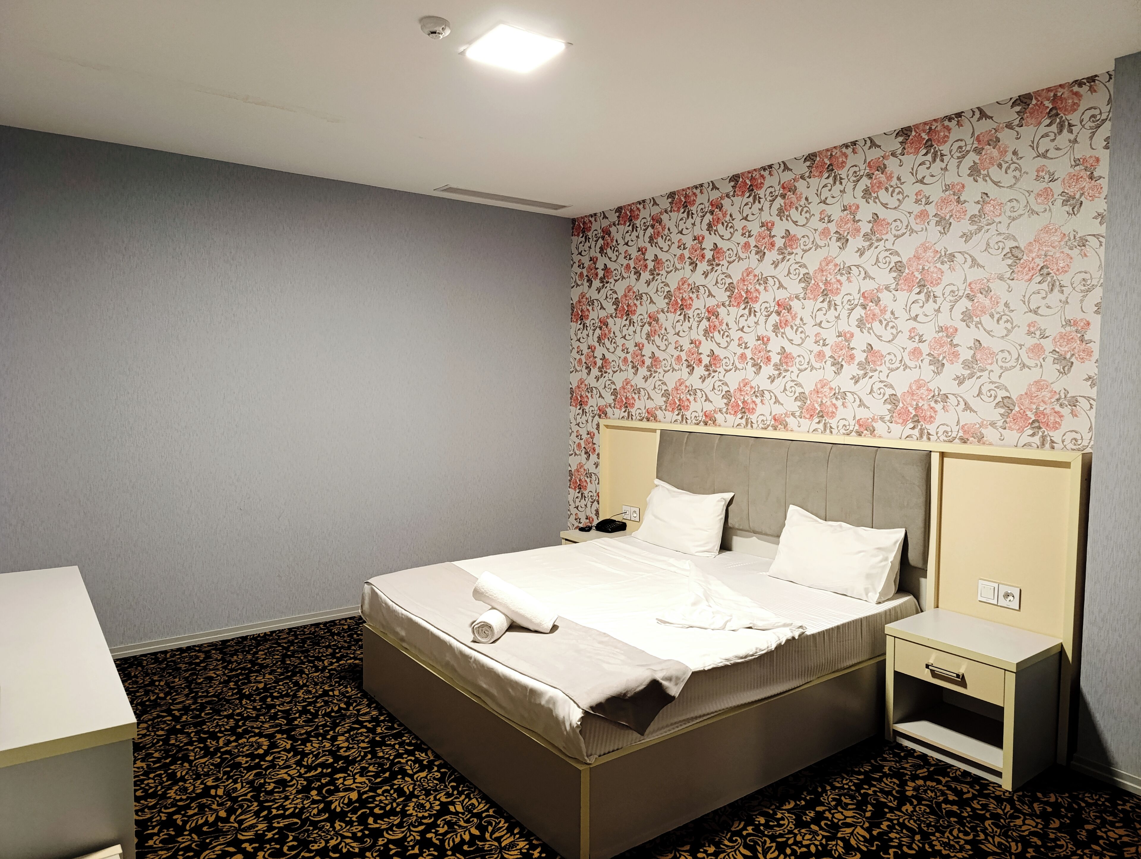 A hotel room with a bed, bedside tables, a desk, and a patterned carpet.