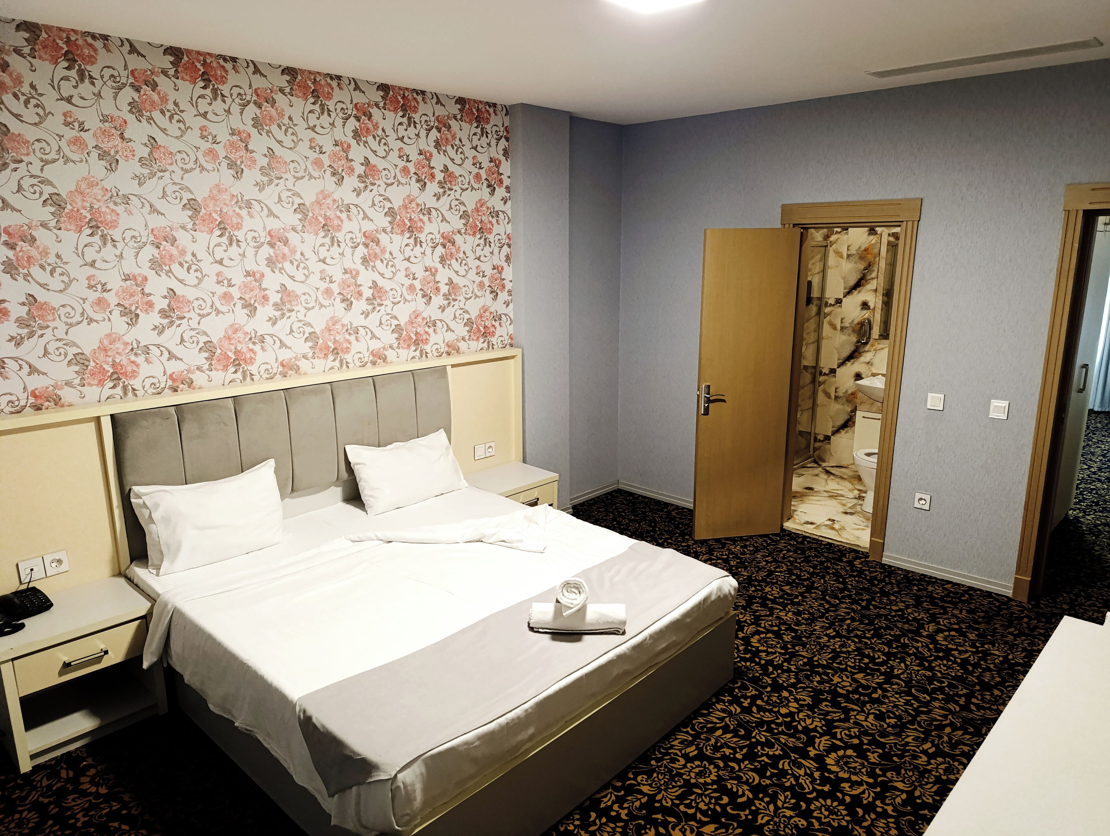 Une chambre d’hôtel avec un lit, une table de chevet, une porte en bois et une salle de bain visible à travers une porte en verre.