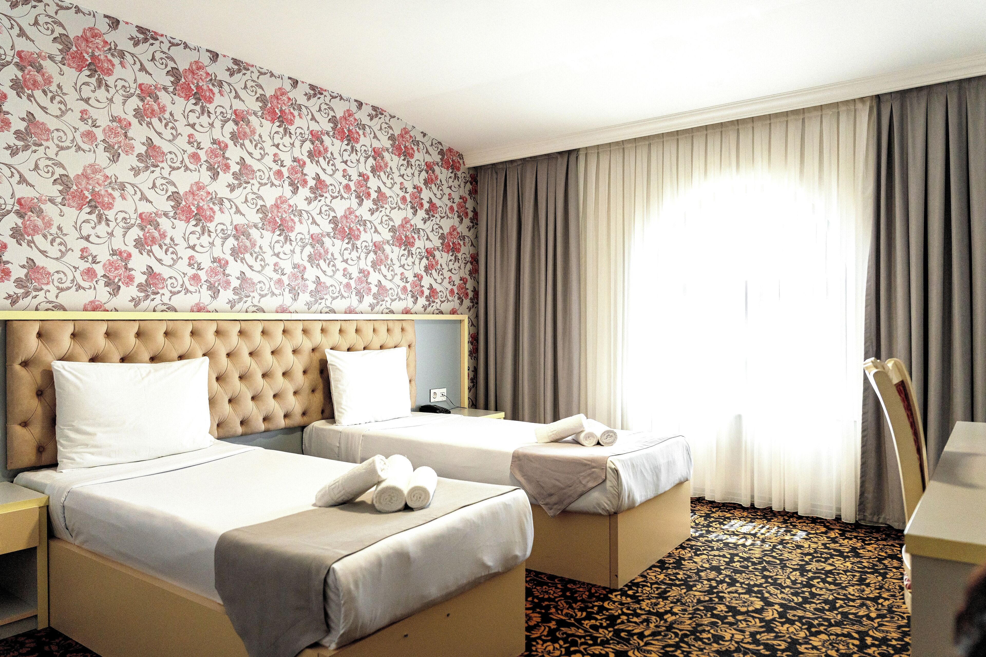 Habitación de hotel con dos camas, cabecera capitonada, papel tapiz floral y un amplio ventanal con cortinas.