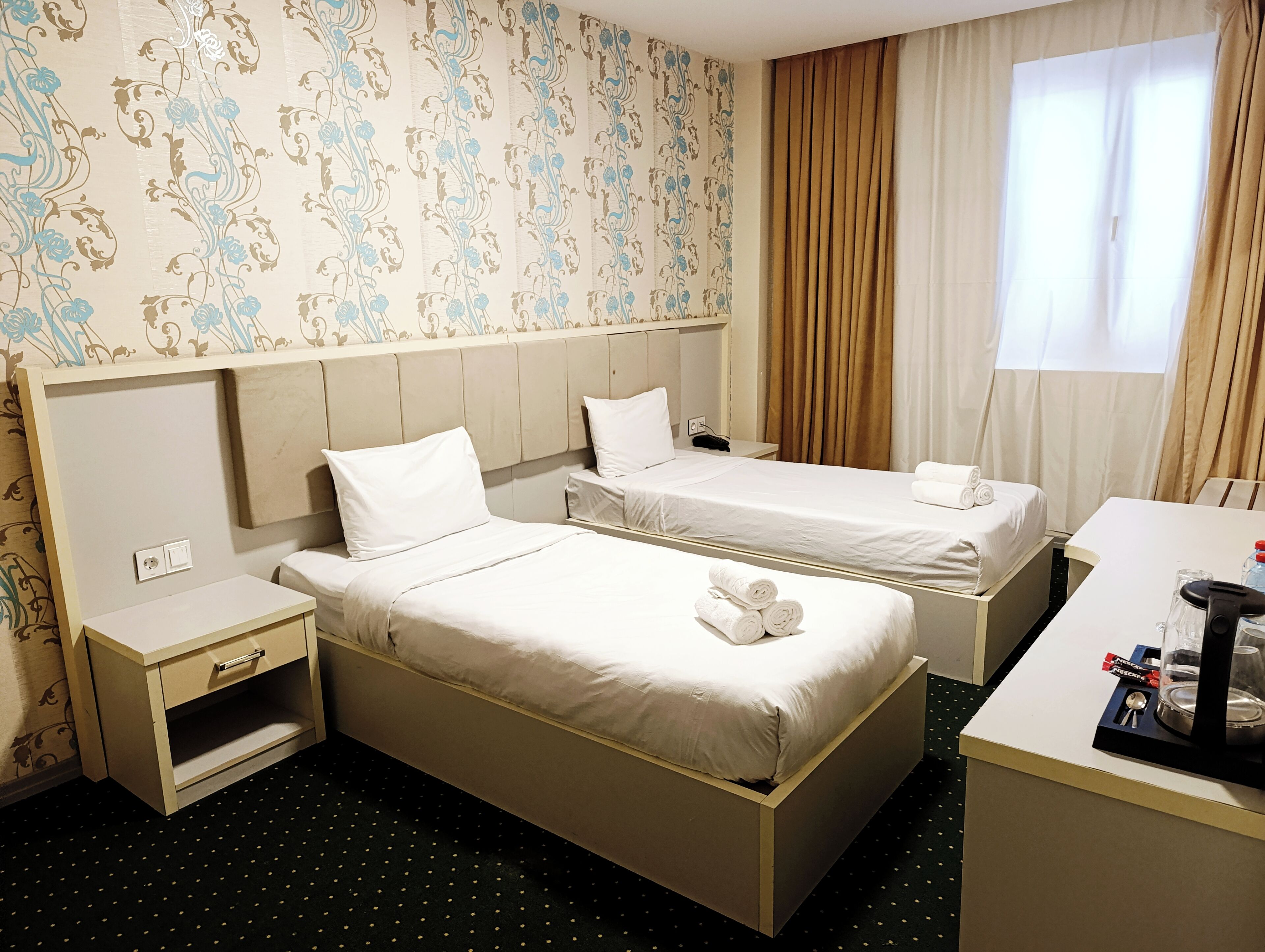 A hotel room with two single beds, a desk with a chair, a nightstand, and a window with curtains.