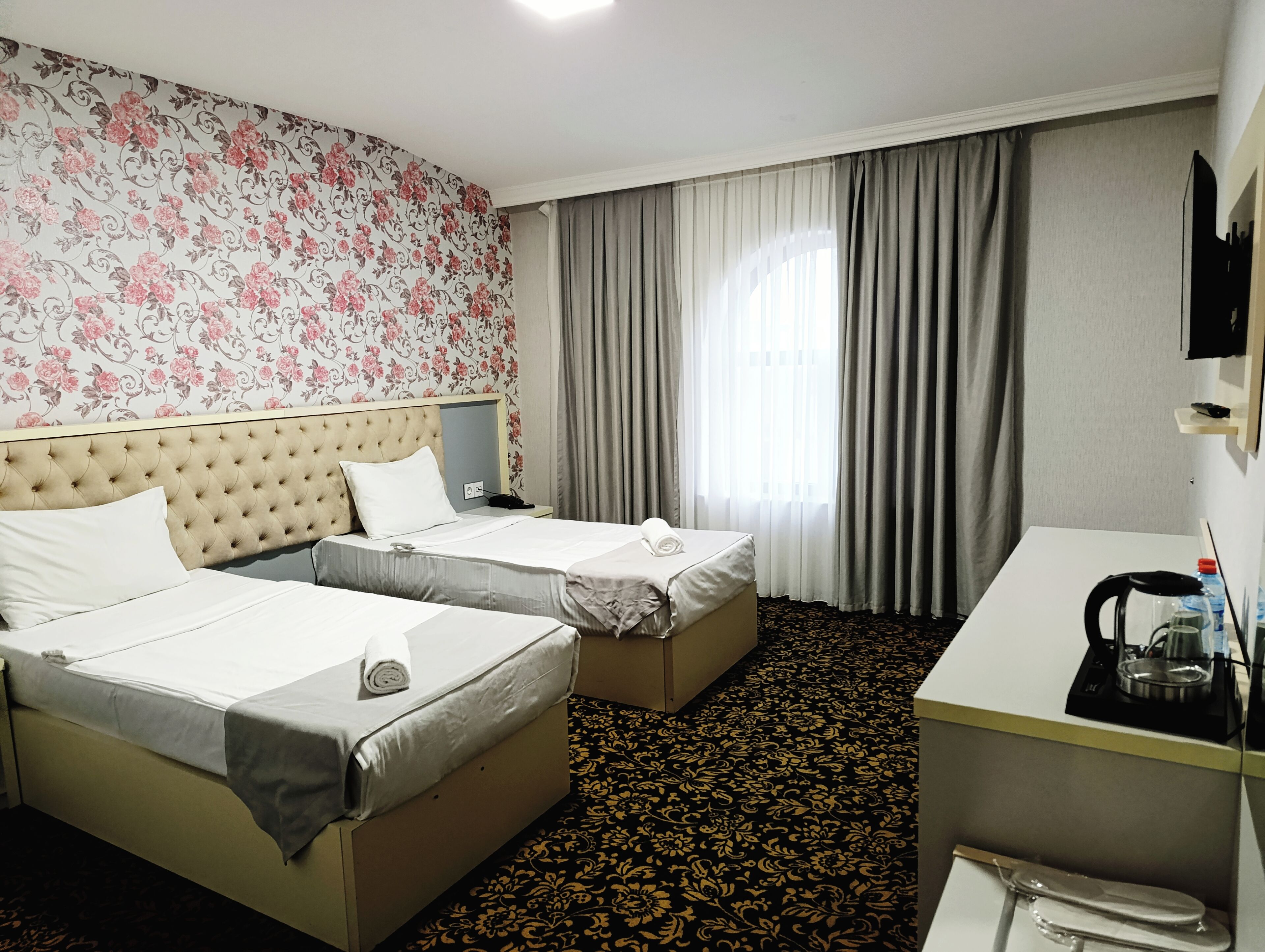 Quarto de hotel com duas camas, uma escrivaninha, uma cadeira, televisão e janela com cortinas.