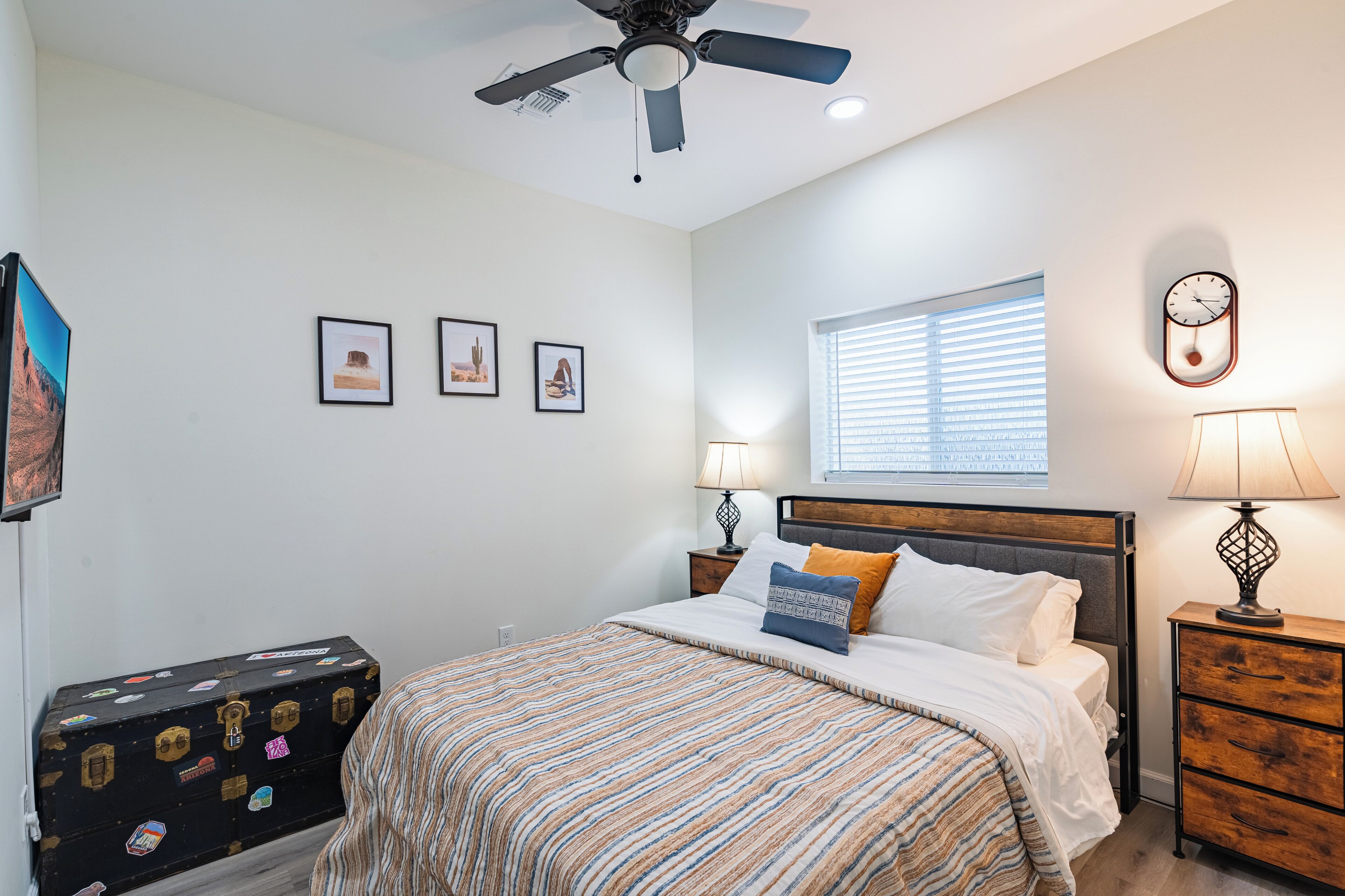 A bedroom with a bed, two bedside tables with lamps, a ceiling fan, a window with blinds, and a wall-mounted clock.