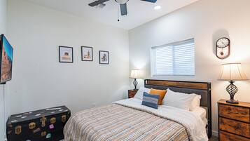 A bedroom with a bed, two bedside tables with lamps, a ceiling fan, a window with blinds, and a wall-mounted clock.