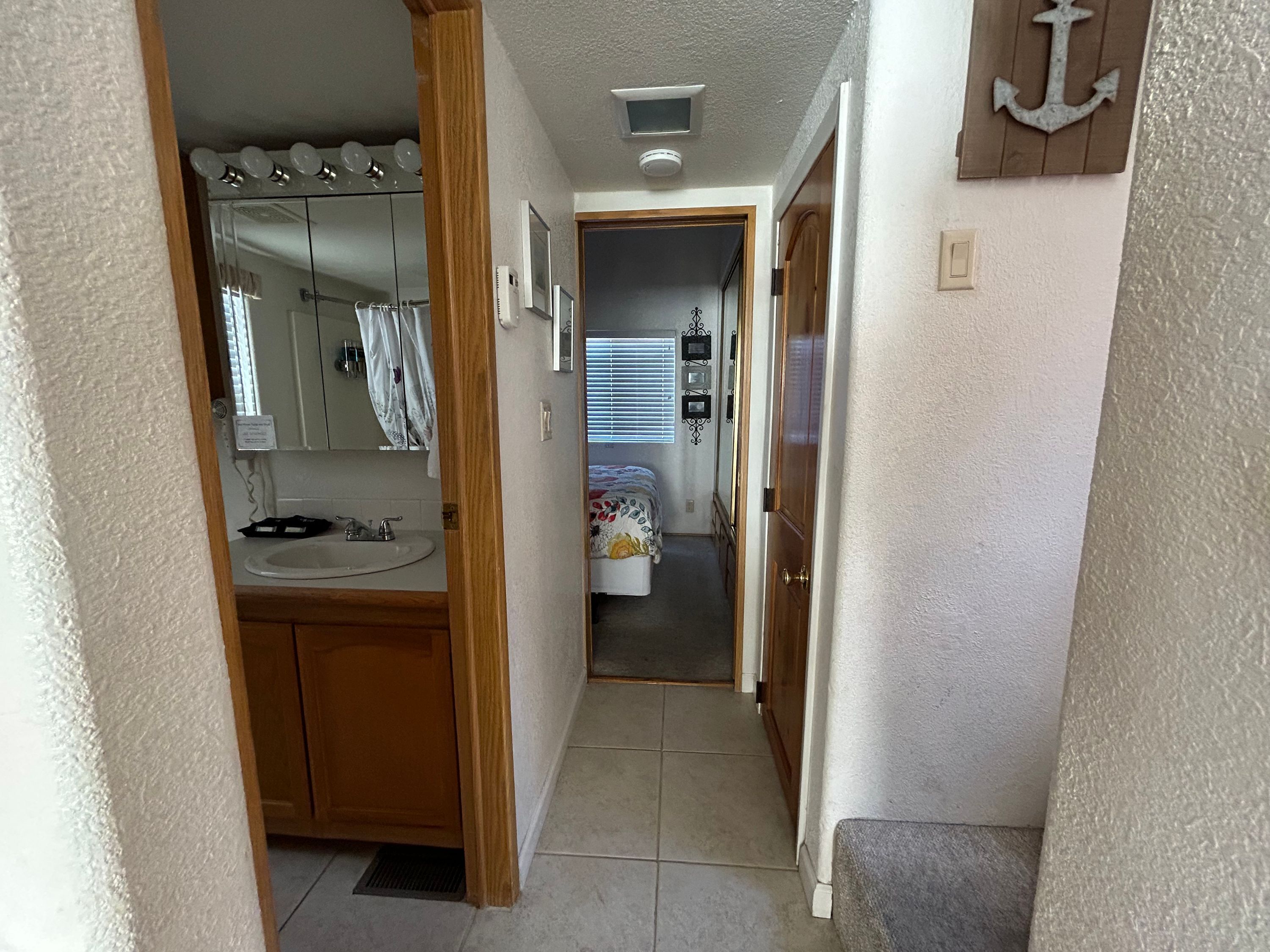 Ein schmaler Flur mit einem Badezimmer links, inklusive Waschbecken, Spiegel und Waschtisch. Am Ende des Flurs ist ein Schlafzimmer mit Bett und Fensterläden zu sehen.