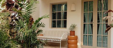 Een terras met een witte metalen bank, een plant in pot en een raam met uitzicht op groen.