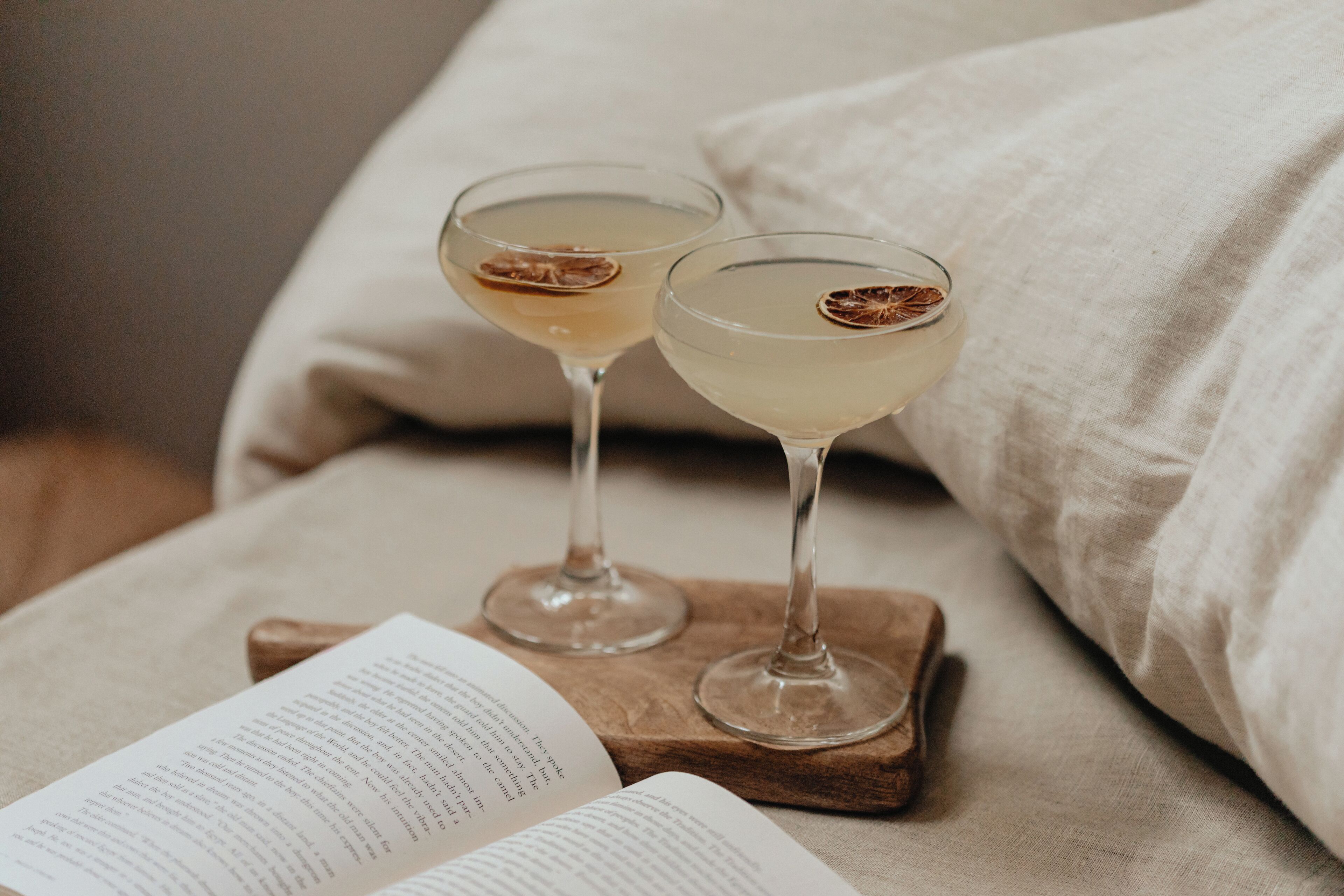 Two glasses of a pale beverage on a wooden coaster with an open book beside them.