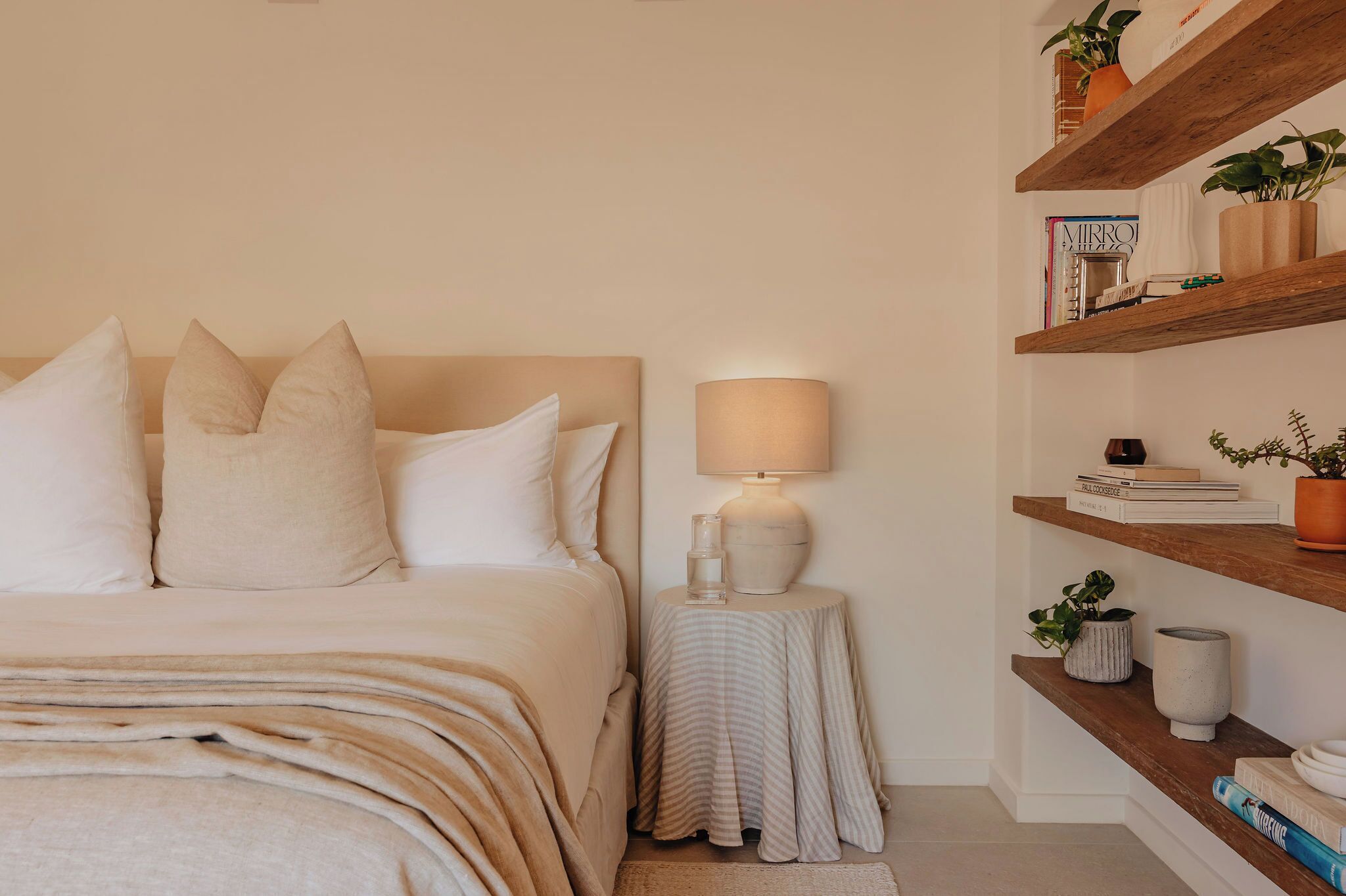 Un dormitorio con cama, mesita de noche, lámpara y estanterías con libros y plantas.