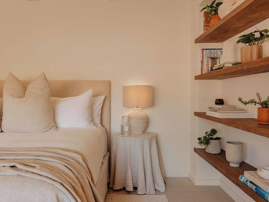 Un dormitorio con cama, mesita de noche, lámpara y estanterías con libros y plantas.