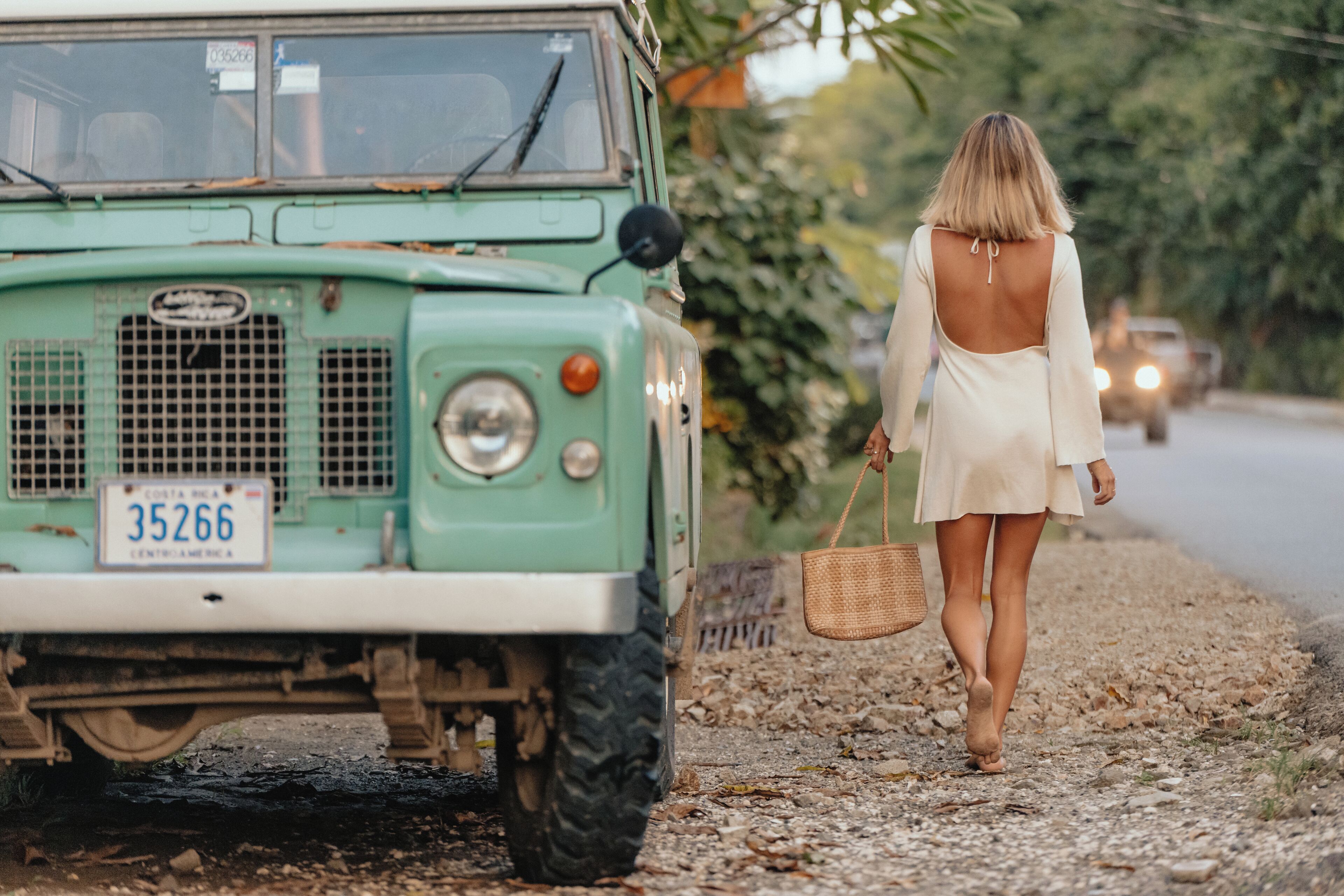 A vintage green vehicle parked on a gravel path with a woman walking beside it.