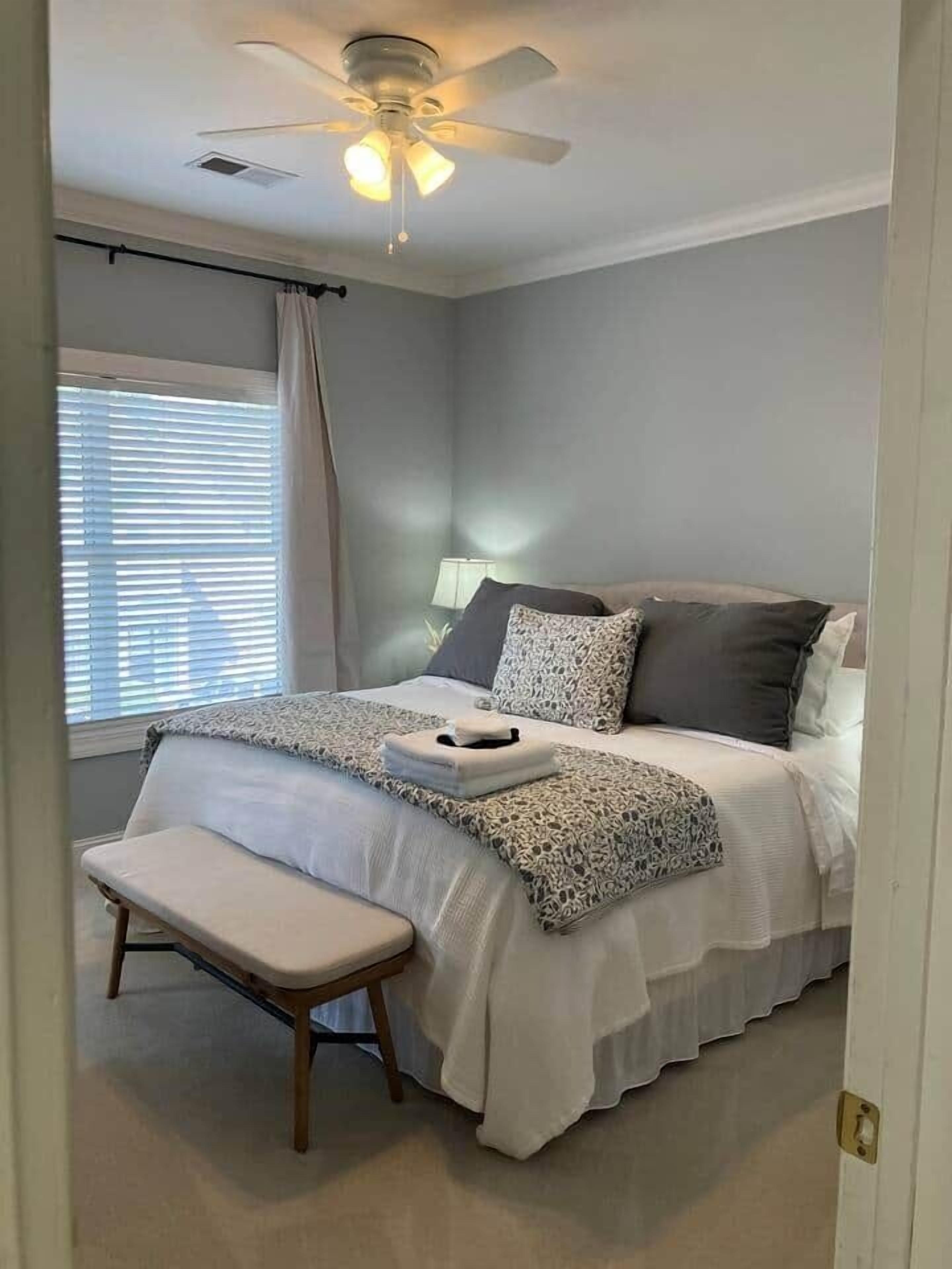 Un dormitorio con cama, mesita de noche, banco, ventilador de techo y ventana con persianas.