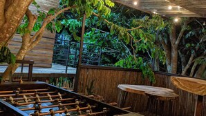 Un espace de loisirs intérieur comprenant une table de baby-foot, des tables et des chaises en bois, le tout entouré d’une végétation luxuriante et surmonté d’un plafond en métal ondulé.