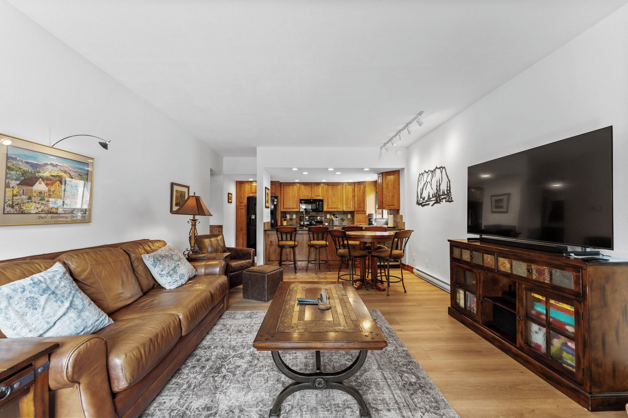 Una sala moderna con un sofá de cuero, una mesita de centro y un televisor sobre un mueble de madera. Se observa una cocina al fondo, con gabinetes de madera, y un comedor con sillas.