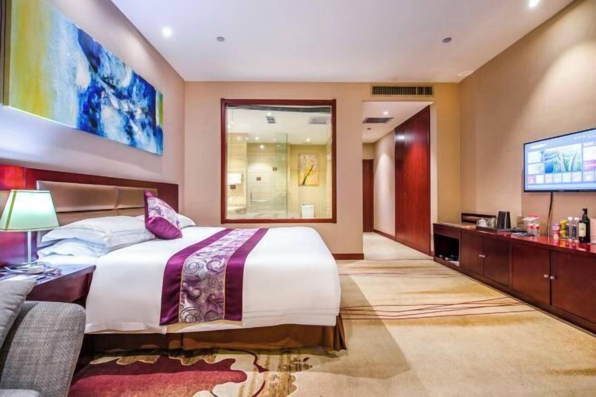 A hotel room with a large bed, a television, a desk, and a bathroom visible through a glass door.