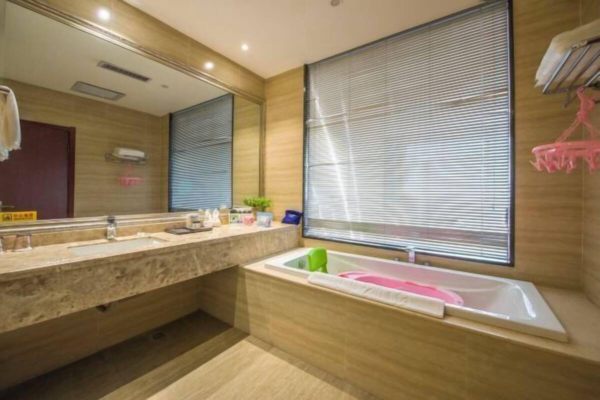 A modern bathroom with a large mirror, two sinks, a bathtub, and a window with blinds.