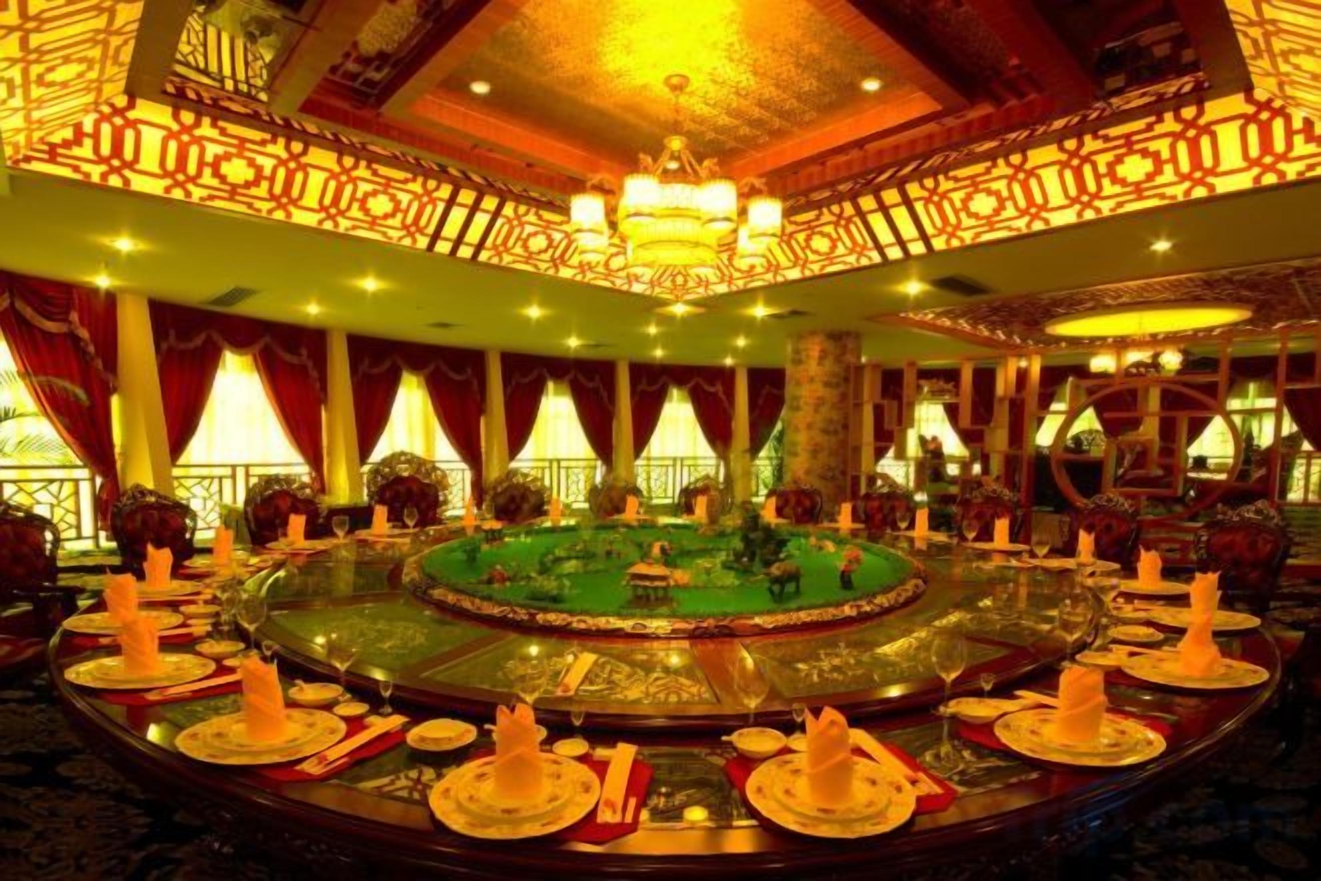Une table de salle à manger ronde, avec un centre décoratif vert, entourée de chaises et de bougies, dans une pièce aux murs à motifs et ornée d’un lustre.
