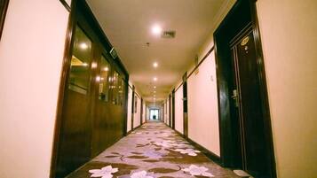 A long, narrow hotel corridor with doors on both sides, a carpet with a floral pattern, and a ceiling with recessed lighting.