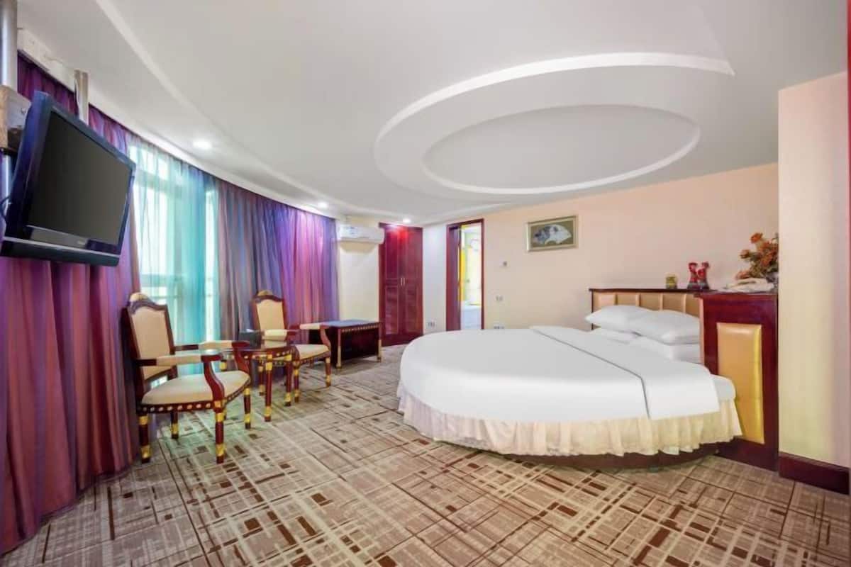Ein Hotelzimmer mit einem großen Bett, einem Schreibtisch mit Stuhl, einem an der Wand befestigten Fernseher und einem gemusterten Teppich.