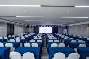 Un salón de conferencias con filas de mesas y sillas, un escenario con una pantalla de proyecciones y una pizarra.