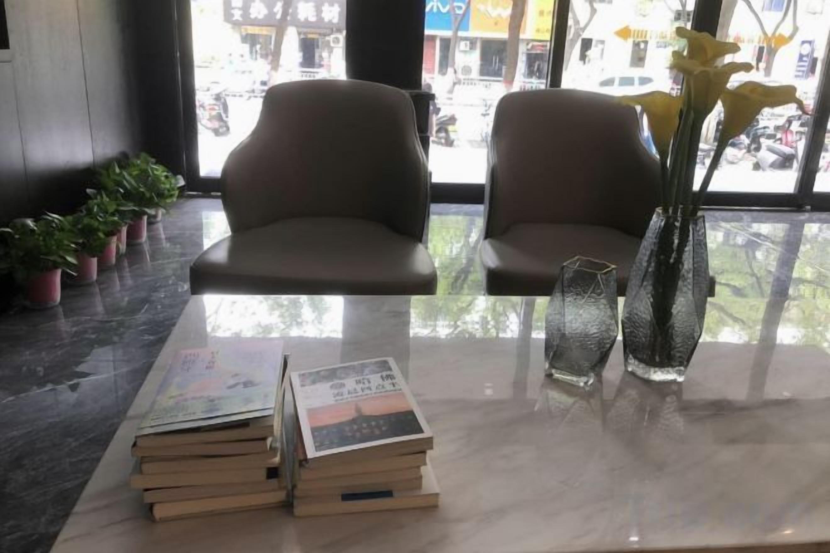 Un área de estar con dos sillas, una mesa con libros y un florero, y vista a la calle a través de una ventana.