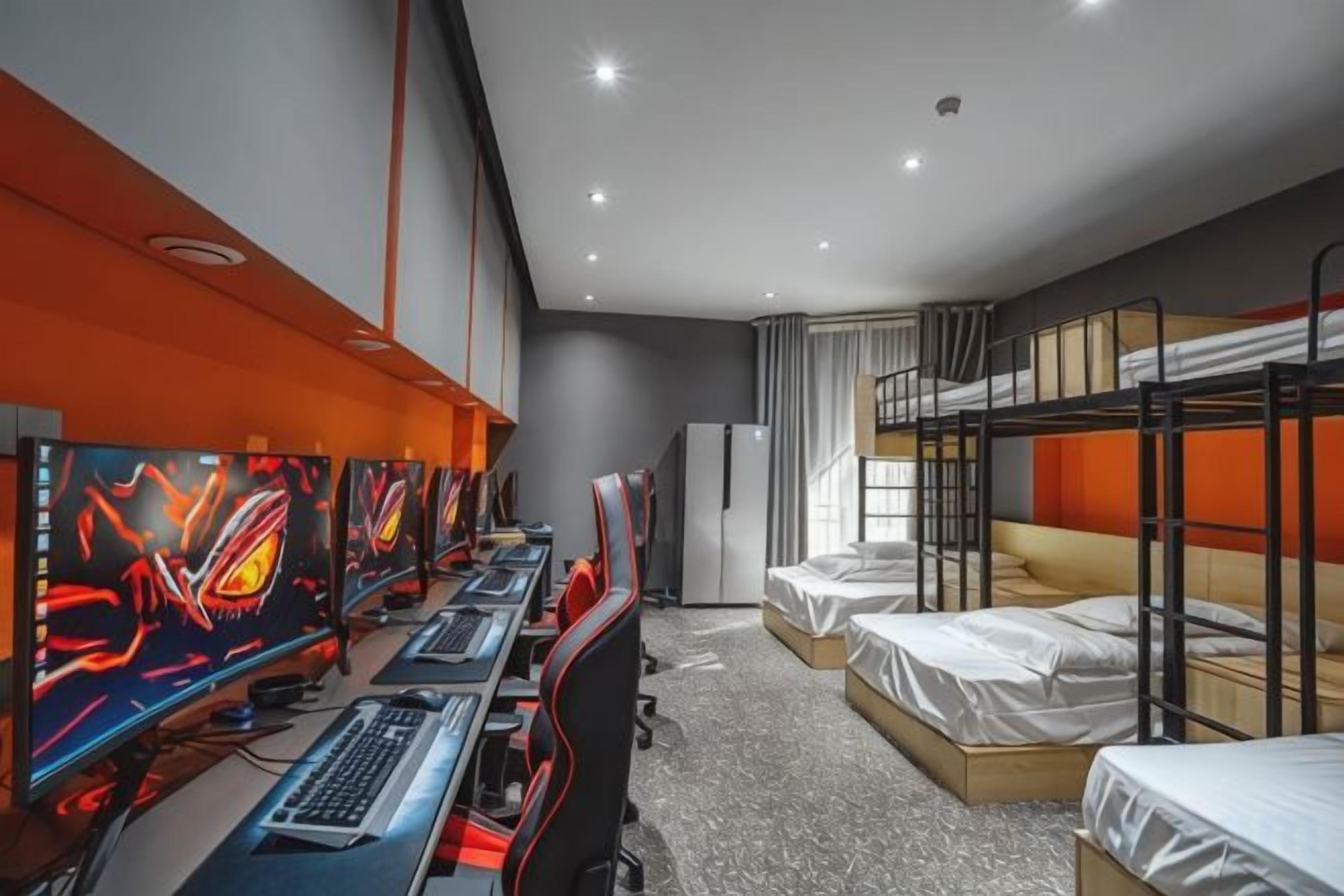 A hotel room with bunk beds, a desk with multiple monitors, and a refrigerator.