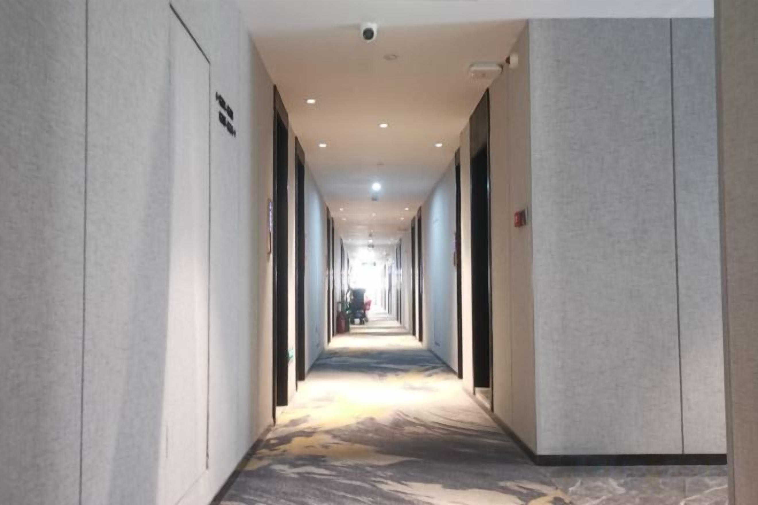 A long, narrow hotel corridor with a patterned carpet, walls with a textured finish, and a ceiling with recessed lighting.
