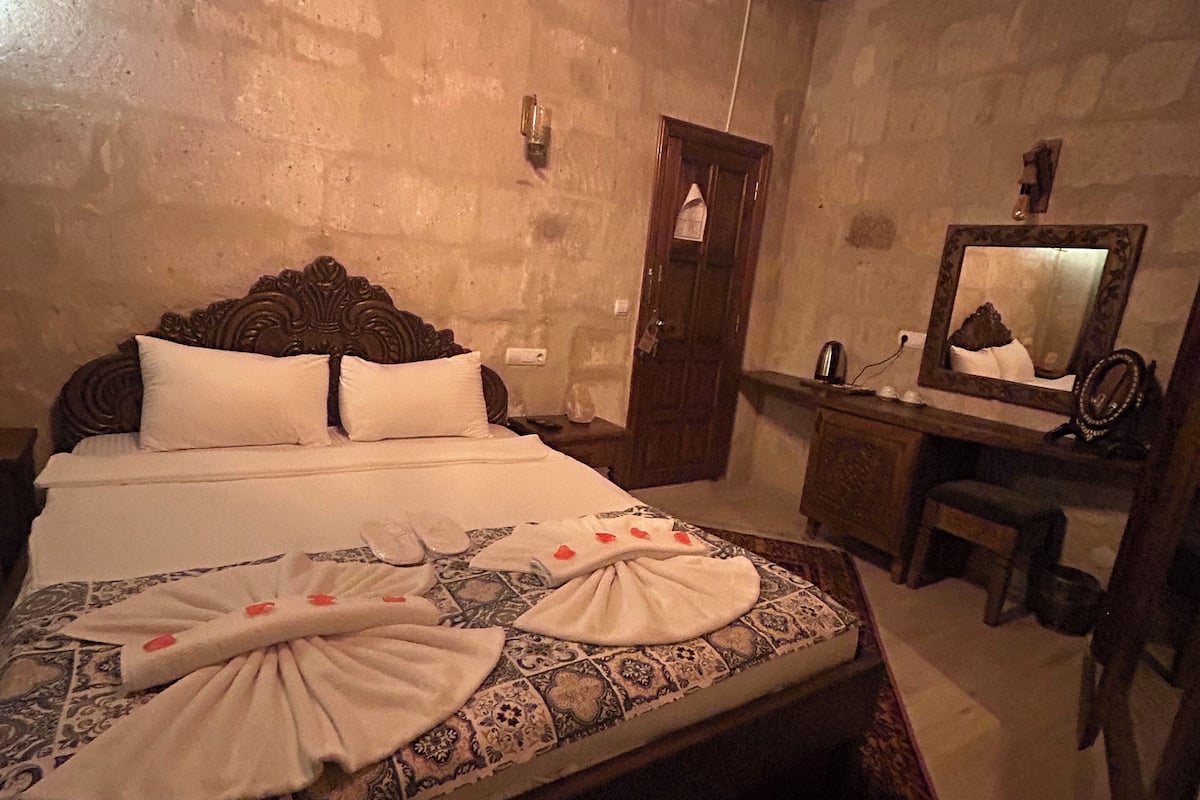 Habitació d'hotel amb un llit, un reposacapses de fusta, un taulell de nit, un mirall i una porta.